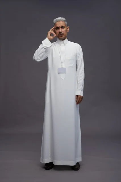 بورتريه لرجل عربي سعودي  خليجي يرتدي الثوب الابيض يقف و يقوم بايماءات واشارات مختلفه امام الكاميرا, خلفيه رماديه , كامل الجسم