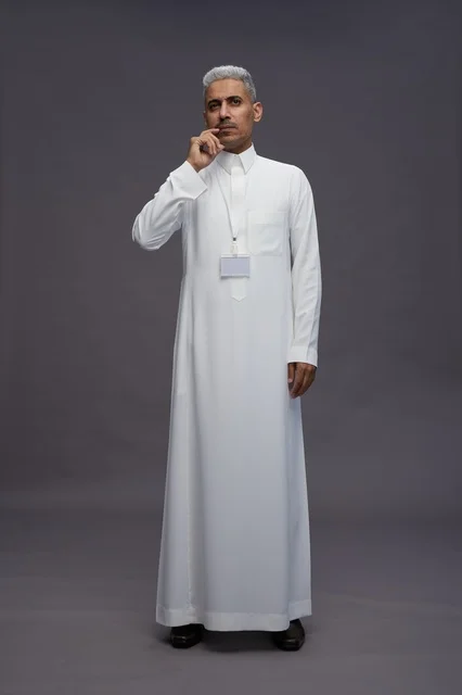 بورتريه لرجل عربي سعودي  خليجي يرتدي الثوب الابيض يقف و يقوم بايماءات واشارات مختلفه امام الكاميرا, خلفيه رماديه , كامل الجسم