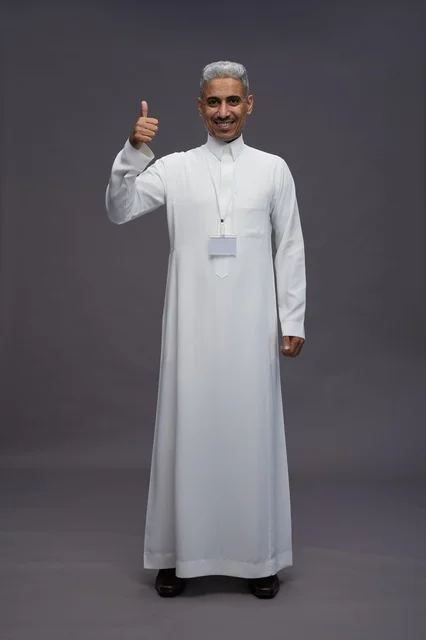 بورتريه لرجل عربي سعودي  خليجي يرتدي الثوب الابيض يقف و يقوم بايماءات واشارات مختلفه امام الكاميرا, خلفيه رماديه , كامل الجسم