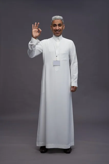 بورتريه لرجل عربي سعودي  خليجي يرتدي الثوب الابيض يقف و يقوم بايماءات واشارات مختلفه امام الكاميرا, خلفيه رماديه , كامل الجسم