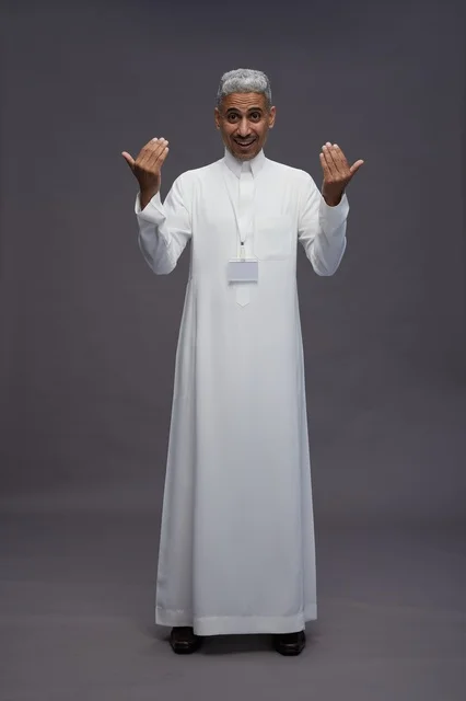 بورتريه لرجل عربي سعودي  خليجي يرتدي الثوب الابيض يقف و يقوم بايماءات واشارات مختلفه امام الكاميرا, خلفيه رماديه , كامل الجسم