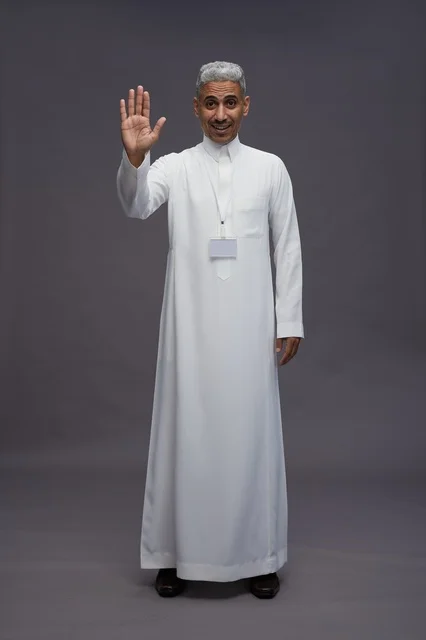 بورتريه لرجل عربي سعودي  خليجي يرتدي الثوب الابيض يقف و يقوم بايماءات واشارات مختلفه امام الكاميرا, خلفيه رماديه , كامل الجسم