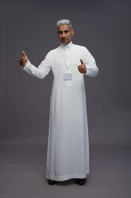 بورتريه لرجل عربي سعودي  خليجي يرتدي الثوب الابيض يقف و يقوم بايماءات واشارات مختلفه امام الكاميرا, خلفيه رماديه , كامل الجسم