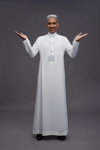 بورتريه لرجل عربي سعودي  خليجي يرتدي الثوب الابيض يقف و يقوم بايماءات واشارات مختلفه امام الكاميرا, خلفيه رماديه , كامل الجسم
