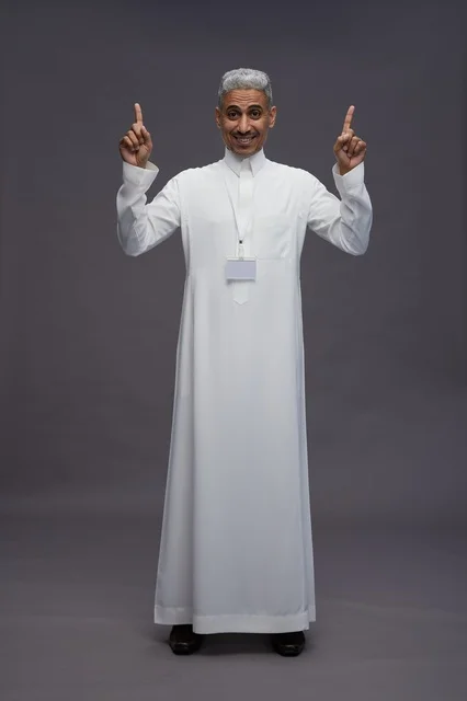 بورتريه لرجل عربي سعودي  خليجي يرتدي الثوب الابيض يقف و يقوم بايماءات واشارات مختلفه امام الكاميرا, خلفيه رماديه , كامل الجسم