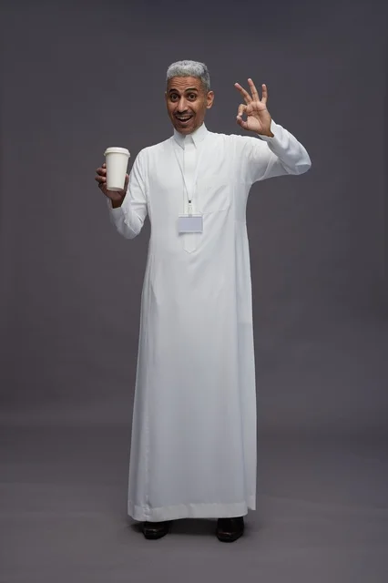 بورتريه لرجل عربي سعودي خليجي يرتدي الثوب الابيض ويستمتع بشرب القهوه, خلفيه رماديه, كامل الجسم