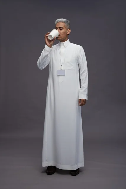 بورتريه لرجل عربي سعودي خليجي يرتدي الثوب الابيض ويستمتع بشرب القهوه, خلفيه رماديه, كامل الجسم