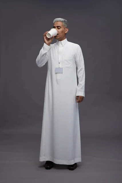 بورتريه لرجل عربي سعودي خليجي يرتدي الثوب الابيض ويستمتع بشرب القهوه, خلفيه رماديه, كامل الجسم