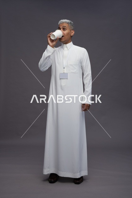 بورتريه لرجل عربي سعودي خليجي يرتدي الثوب الابيض ويستمتع بشرب القهوه, خلفيه رماديه, كامل الجسم