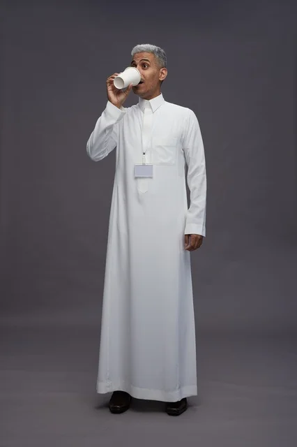 بورتريه لرجل عربي سعودي خليجي يرتدي الثوب الابيض ويستمتع بشرب القهوه, خلفيه رماديه, كامل الجسم