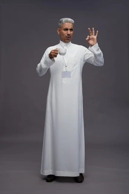 A portrait of an Arab Saudi Gulf man wearing a white thobe and enjoying drinking coffee, with a gray background, full body.