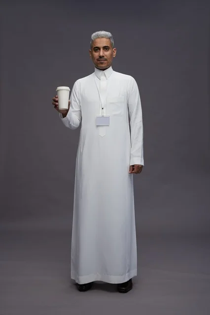 بورتريه لرجل عربي سعودي خليجي يرتدي الثوب الابيض ويستمتع بشرب القهوه, خلفيه رماديه, كامل الجسم
