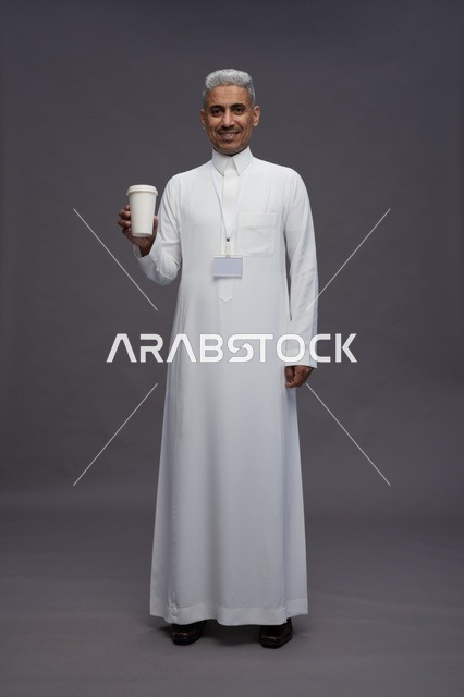 بورتريه لرجل عربي سعودي خليجي يرتدي الثوب الابيض ويستمتع بشرب القهوه, خلفيه رماديه, كامل الجسم