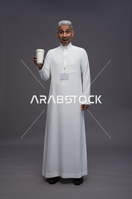 بورتريه لرجل عربي سعودي خليجي يرتدي الثوب الابيض ويستمتع بشرب القهوه, خلفيه رماديه, كامل الجسم