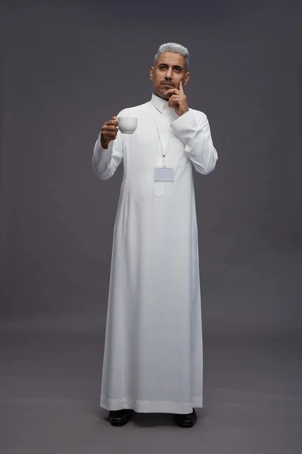 بورتريه لرجل عربي سعودي خليجي يرتدي الثوب الابيض ويستمتع بشرب القهوه, خلفيه رماديه, كامل الجسم