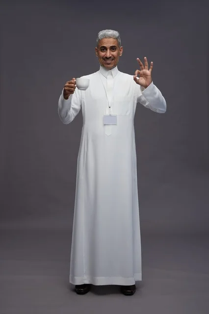بورتريه لرجل عربي سعودي خليجي يرتدي الثوب الابيض ويستمتع بشرب القهوه, خلفيه رماديه, كامل الجسم