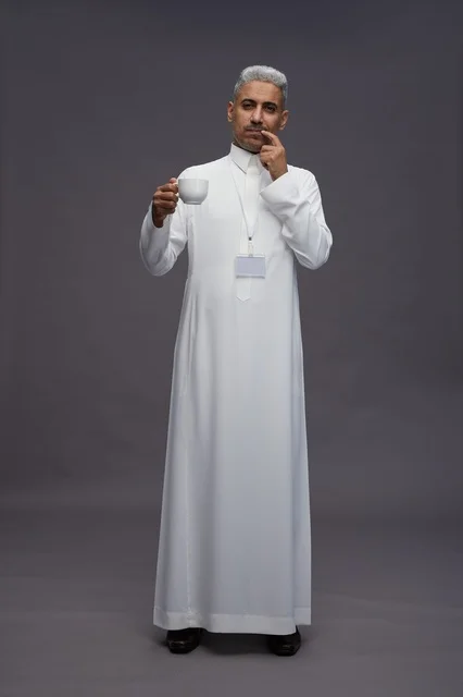 بورتريه لرجل عربي سعودي خليجي يرتدي الثوب الابيض ويستمتع بشرب القهوه, خلفيه رماديه, كامل الجسم