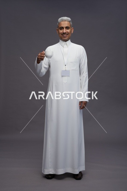 بورتريه لرجل عربي سعودي خليجي يرتدي الثوب الابيض ويستمتع بشرب القهوه, خلفيه رماديه, كامل الجسم