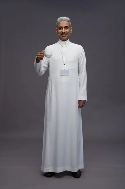 بورتريه لرجل عربي سعودي خليجي يرتدي الثوب الابيض ويستمتع بشرب القهوه, خلفيه رماديه, كامل الجسم