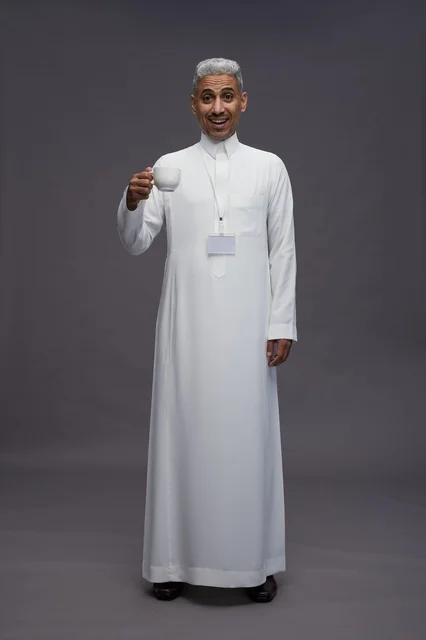 بورتريه لرجل عربي سعودي خليجي يرتدي الثوب الابيض ويستمتع بشرب القهوه, خلفيه رماديه, كامل الجسم