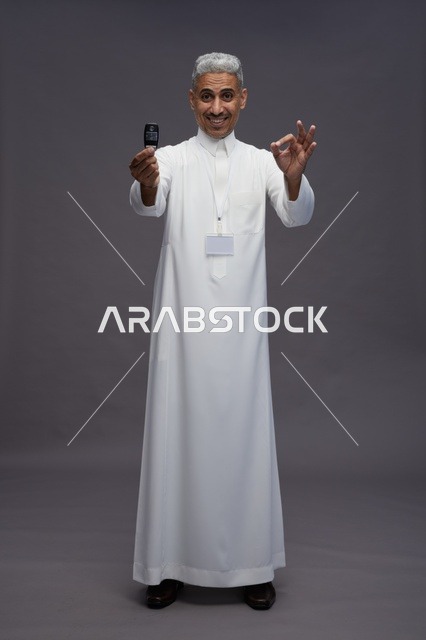 رجل عربي  سعودي  خليجي يرتدي الثوب الابيض يحمل مفتاح سيارة, مفهوم امتلاك سياره جديده, خلفيه رماديه