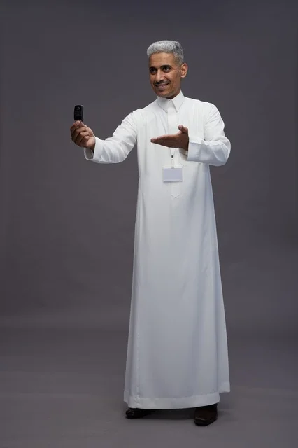 رجل عربي  سعودي  خليجي يرتدي الثوب الابيض يحمل مفتاح سيارة, مفهوم امتلاك سياره جديده, خلفيه رماديه
