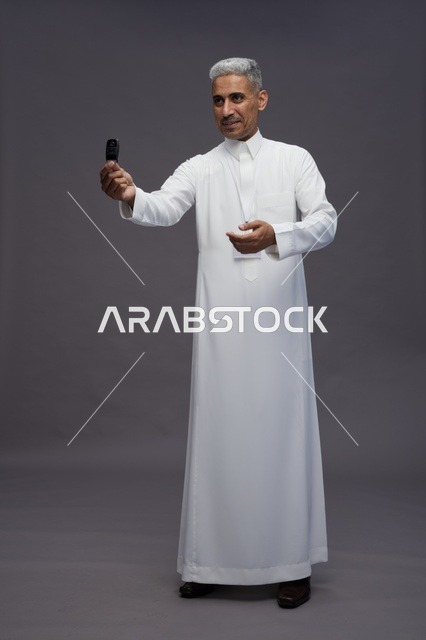 رجل عربي  سعودي  خليجي يرتدي الثوب الابيض يحمل مفتاح سيارة, مفهوم امتلاك سياره جديده, خلفيه رماديه