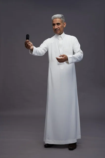 رجل عربي  سعودي  خليجي يرتدي الثوب الابيض يحمل مفتاح سيارة, مفهوم امتلاك سياره جديده, خلفيه رماديه