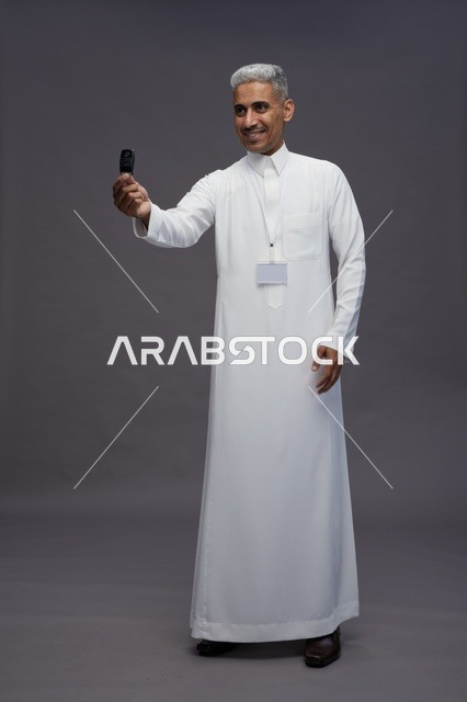 رجل عربي  سعودي  خليجي يرتدي الثوب الابيض يحمل مفتاح سيارة, مفهوم امتلاك سياره جديده, خلفيه رماديه
