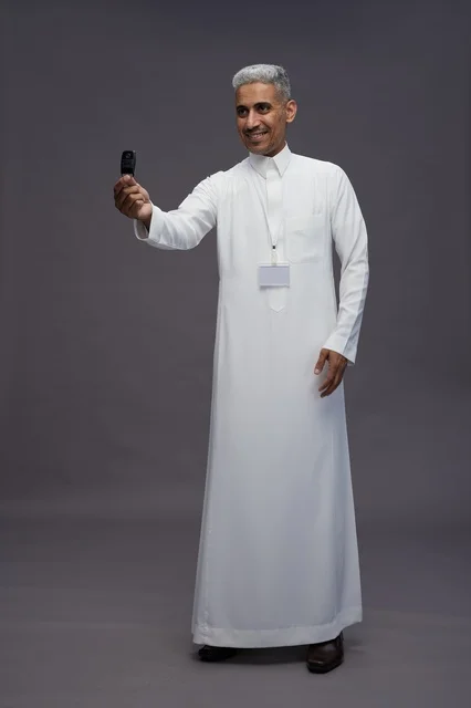 رجل عربي  سعودي  خليجي يرتدي الثوب الابيض يحمل مفتاح سيارة, مفهوم امتلاك سياره جديده, خلفيه رماديه