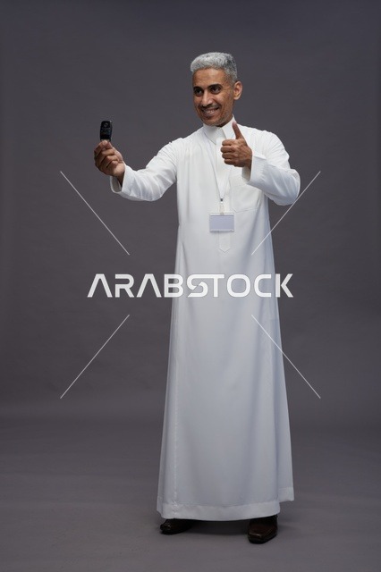 رجل عربي  سعودي  خليجي يرتدي الثوب الابيض يحمل مفتاح سيارة, مفهوم امتلاك سياره جديده, خلفيه رماديه