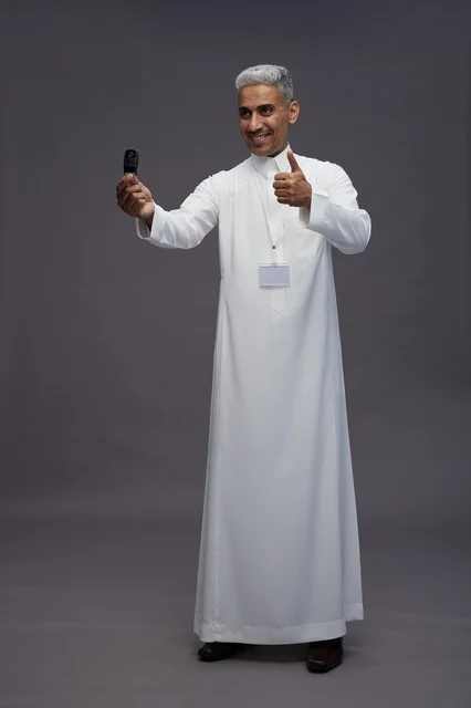 رجل عربي  سعودي  خليجي يرتدي الثوب الابيض يحمل مفتاح سيارة, مفهوم امتلاك سياره جديده, خلفيه رماديه