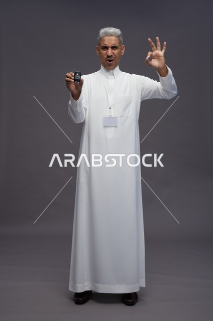 رجل عربي  سعودي  خليجي يرتدي الثوب الابيض يحمل مفتاح سيارة, مفهوم امتلاك سياره جديده, خلفيه رماديه