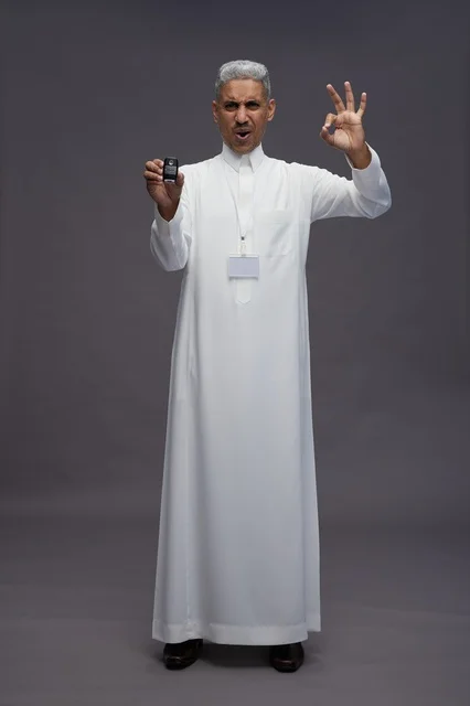 رجل عربي  سعودي  خليجي يرتدي الثوب الابيض يحمل مفتاح سيارة, مفهوم امتلاك سياره جديده, خلفيه رماديه
