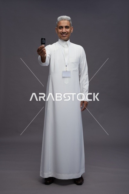 رجل عربي  سعودي  خليجي يرتدي الثوب الابيض يحمل مفتاح سيارة, مفهوم امتلاك سياره جديده, خلفيه رماديه