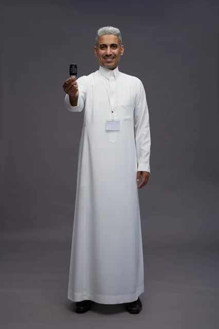 رجل عربي  سعودي  خليجي يرتدي الثوب الابيض يحمل مفتاح سيارة, مفهوم امتلاك سياره جديده, خلفيه رماديه