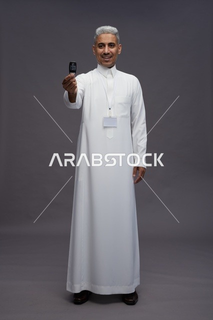 رجل عربي  سعودي  خليجي يرتدي الثوب الابيض يحمل مفتاح سيارة, مفهوم امتلاك سياره جديده, خلفيه رماديه