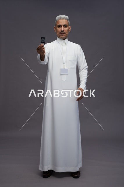 رجل عربي  سعودي  خليجي يرتدي الثوب الابيض يحمل مفتاح سيارة, مفهوم امتلاك سياره جديده, خلفيه رماديه