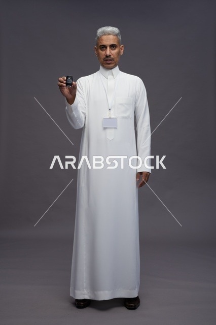 رجل عربي  سعودي  خليجي يرتدي الثوب الابيض يحمل مفتاح سيارة, مفهوم امتلاك سياره جديده, خلفيه رماديه