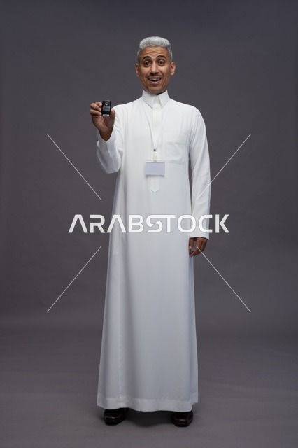 رجل عربي  سعودي  خليجي يرتدي الثوب الابيض يحمل مفتاح سيارة, مفهوم امتلاك سياره جديده, خلفيه رماديه