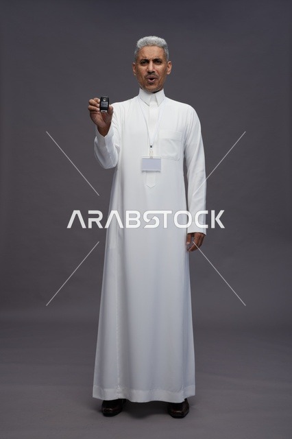 رجل عربي  سعودي  خليجي يرتدي الثوب الابيض يحمل مفتاح سيارة, مفهوم امتلاك سياره جديده, خلفيه رماديه