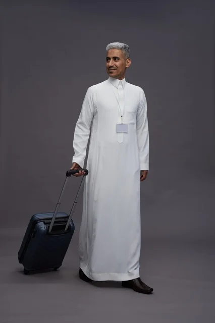 Portrait of a Saudi Arabian Gulf man standing with a suitcase, concept of tourism and travel, gray background, full body.