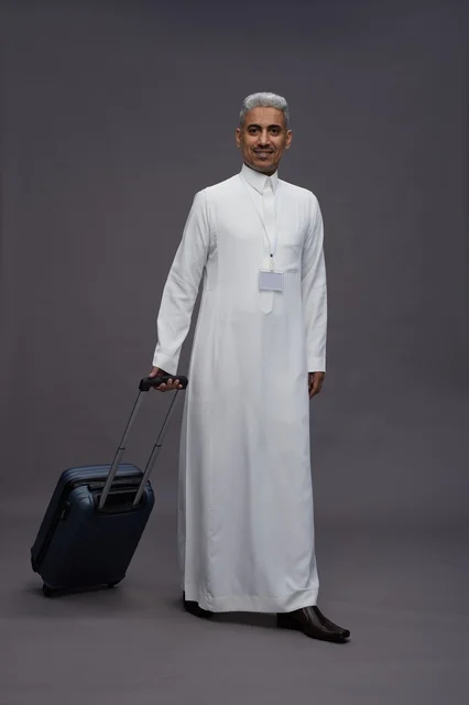 Portrait of a Saudi Arabian Gulf man standing with a suitcase, concept of tourism and travel, gray background, full body.