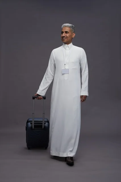 Portrait of a Saudi Arabian Gulf man standing with a suitcase, concept of tourism and travel, gray background, full body.