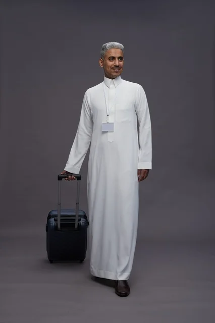 Portrait of a Saudi Arabian Gulf man standing with a suitcase, concept of tourism and travel, gray background, full body.