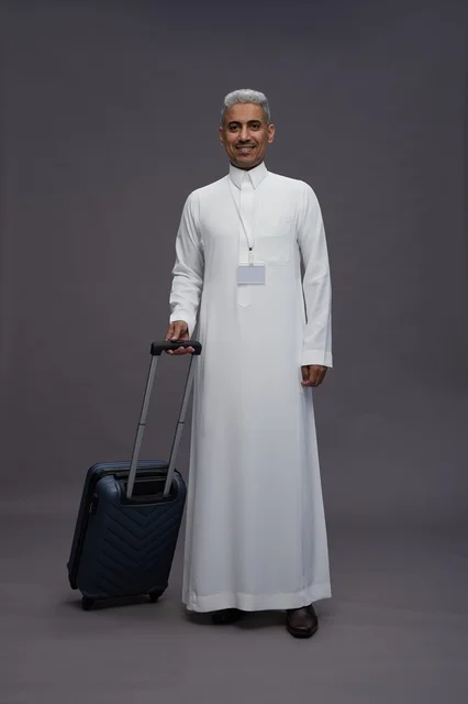 Portrait of a Saudi Arabian Gulf man standing with a suitcase, concept of tourism and travel, gray background, full body.