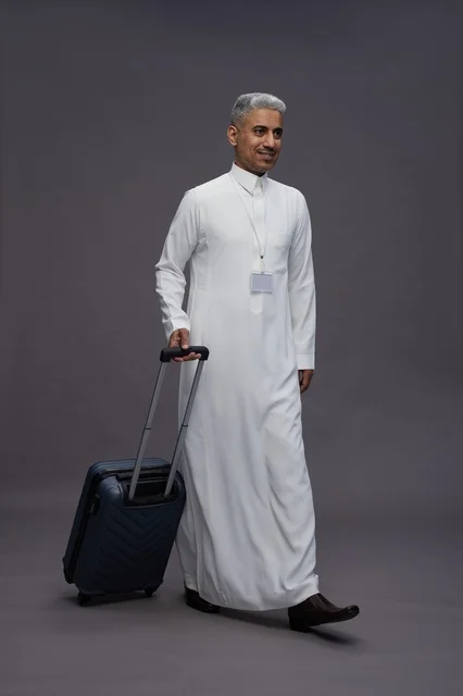 Portrait of a Saudi Arabian Gulf man standing with a suitcase, concept of tourism and travel, gray background, full body.