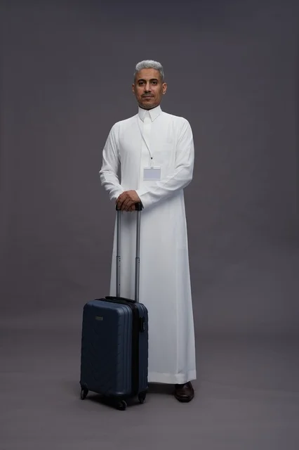 Portrait of a Saudi Arabian Gulf man standing with a suitcase, concept of tourism and travel, gray background, full body.