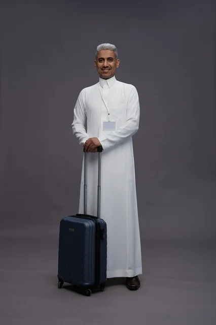 Portrait of a Saudi Arabian Gulf man standing with a suitcase, concept of tourism and travel, gray background, full body.