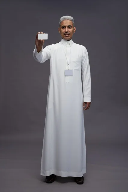 A portrait of an Arab Saudi Gulf man wearing a white thobe displaying a blank white credit card and pointing to it, a personal identification card, full body, gray background.